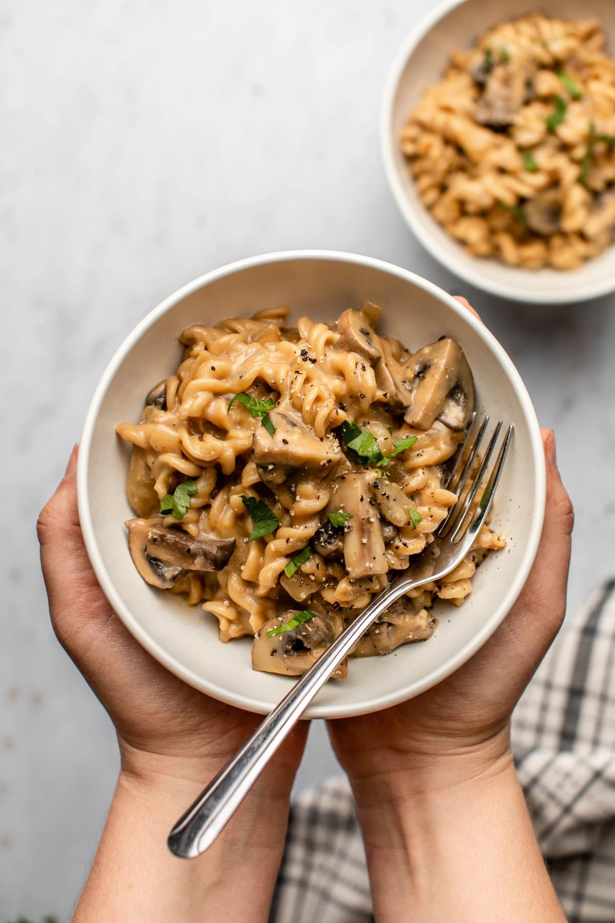 hands holding white bowl of mushroom stroganoff topped with parsley and black pepper
