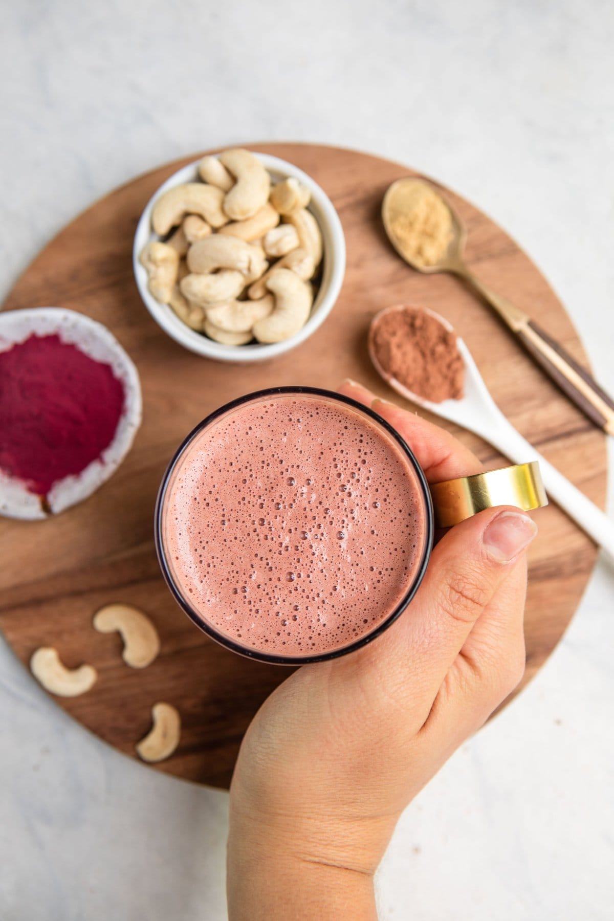 hand holding mug of beet latte with ingredients in background on round wood cutting board