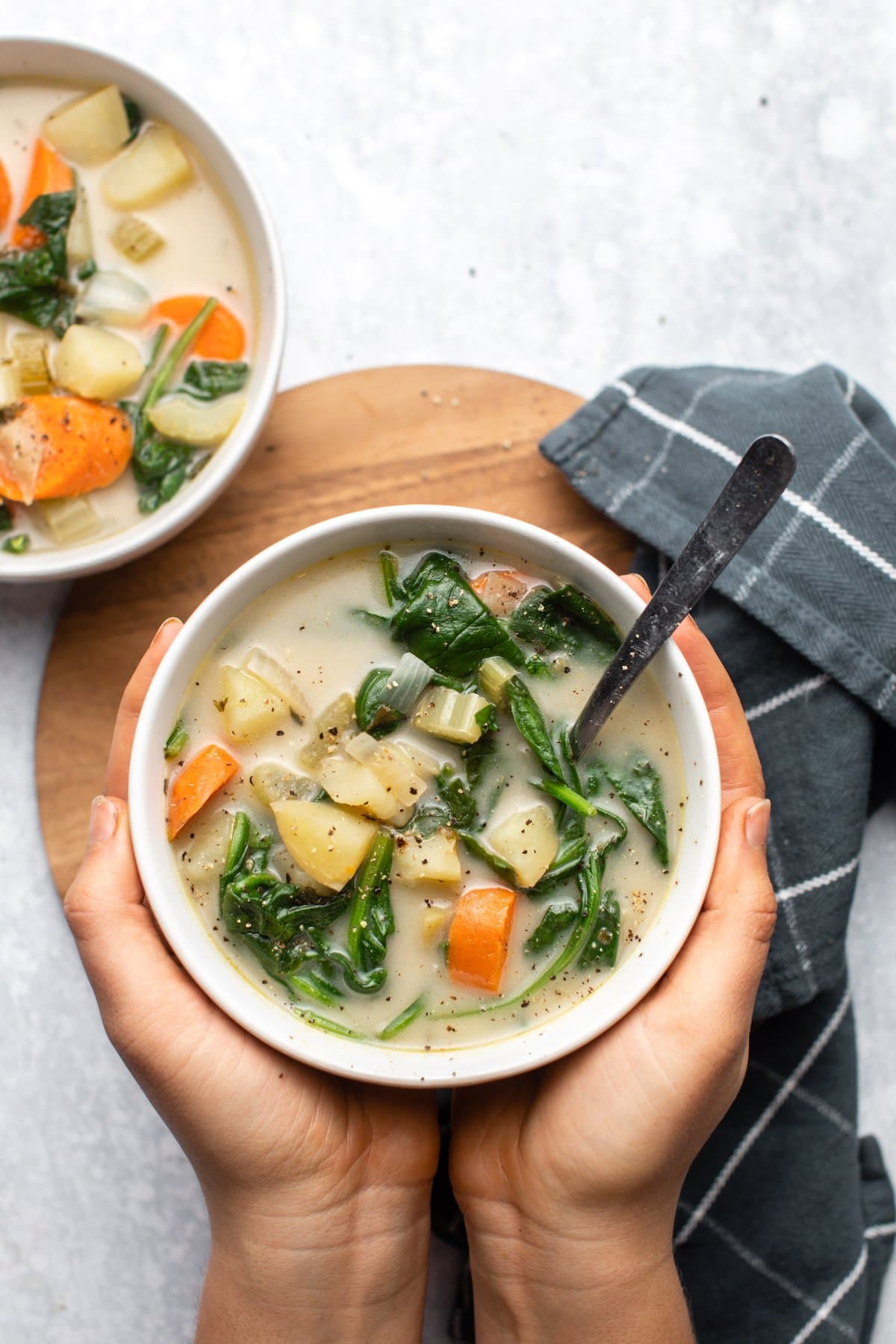 hands holding white bowl of potato spinach soup over wooden serving tray