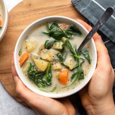 hands holding white bowl of potato spinach soup with silver spoon over wooden serving board