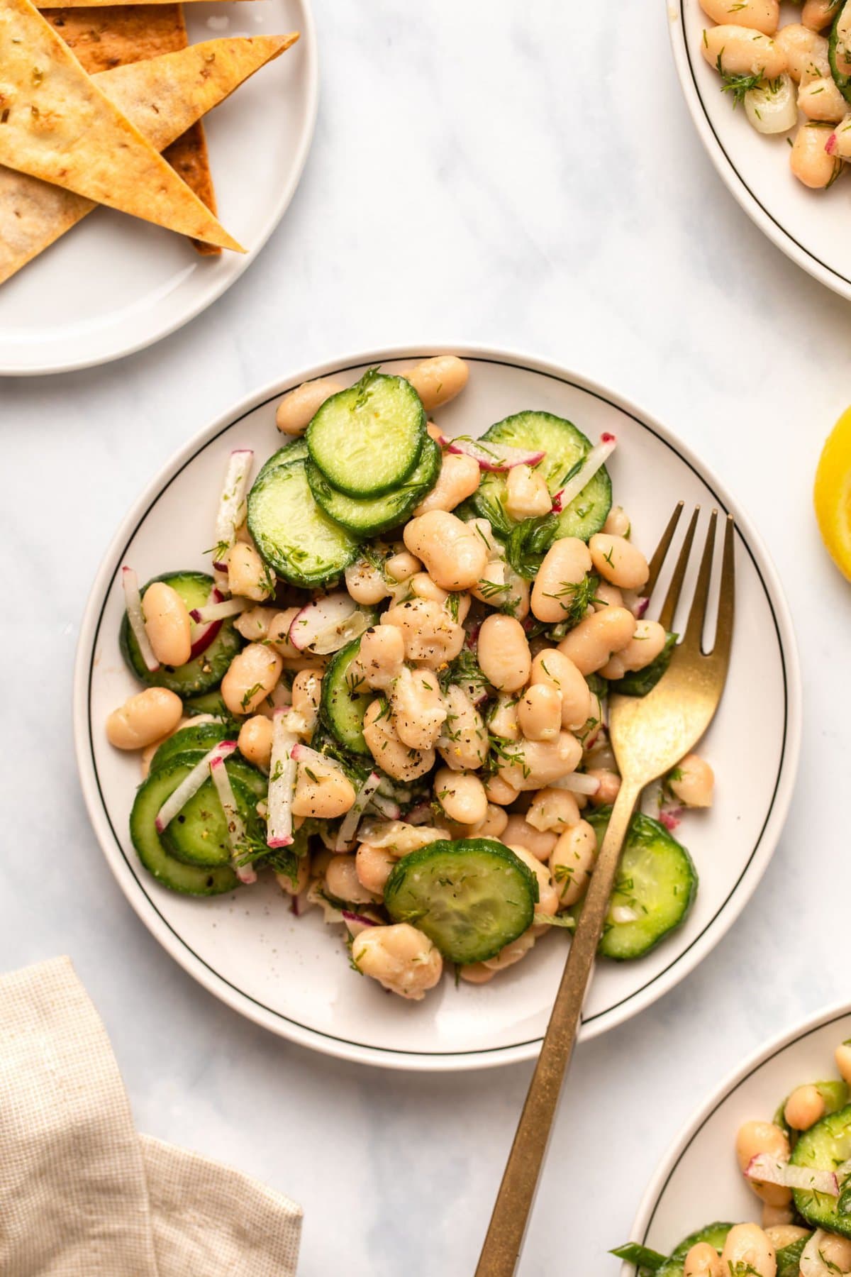 Dilly white bean salad on small white plate with gold fork. A plate of pita chips is in the background