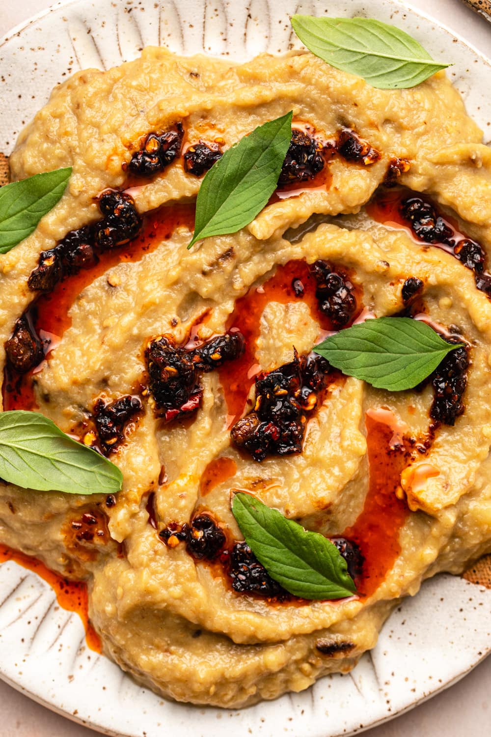 a zoomed in image of garlic miso eggplant dip served on a white plate, topped with chili crisp and thai basil