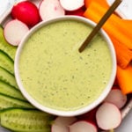 Green Goddess Dressing in small white bowl on plate surrounded by sliced radishes, cucumbers, and carrot
