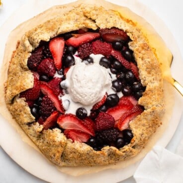 Berry galette topped with melted coco whip on marble serving tray. Fresh flowers lay next to the galette on a white background