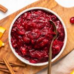 Bowl of cranberry sauce with gold serving spoon on wooden serving board surrounded by orange sliced and cinnamon sticks