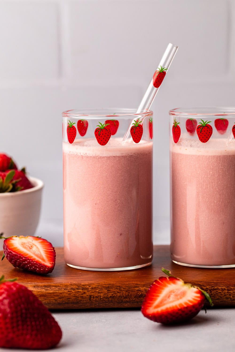 two glasses of strawberry milk served on a cutting board with decorations of strawberry pieces