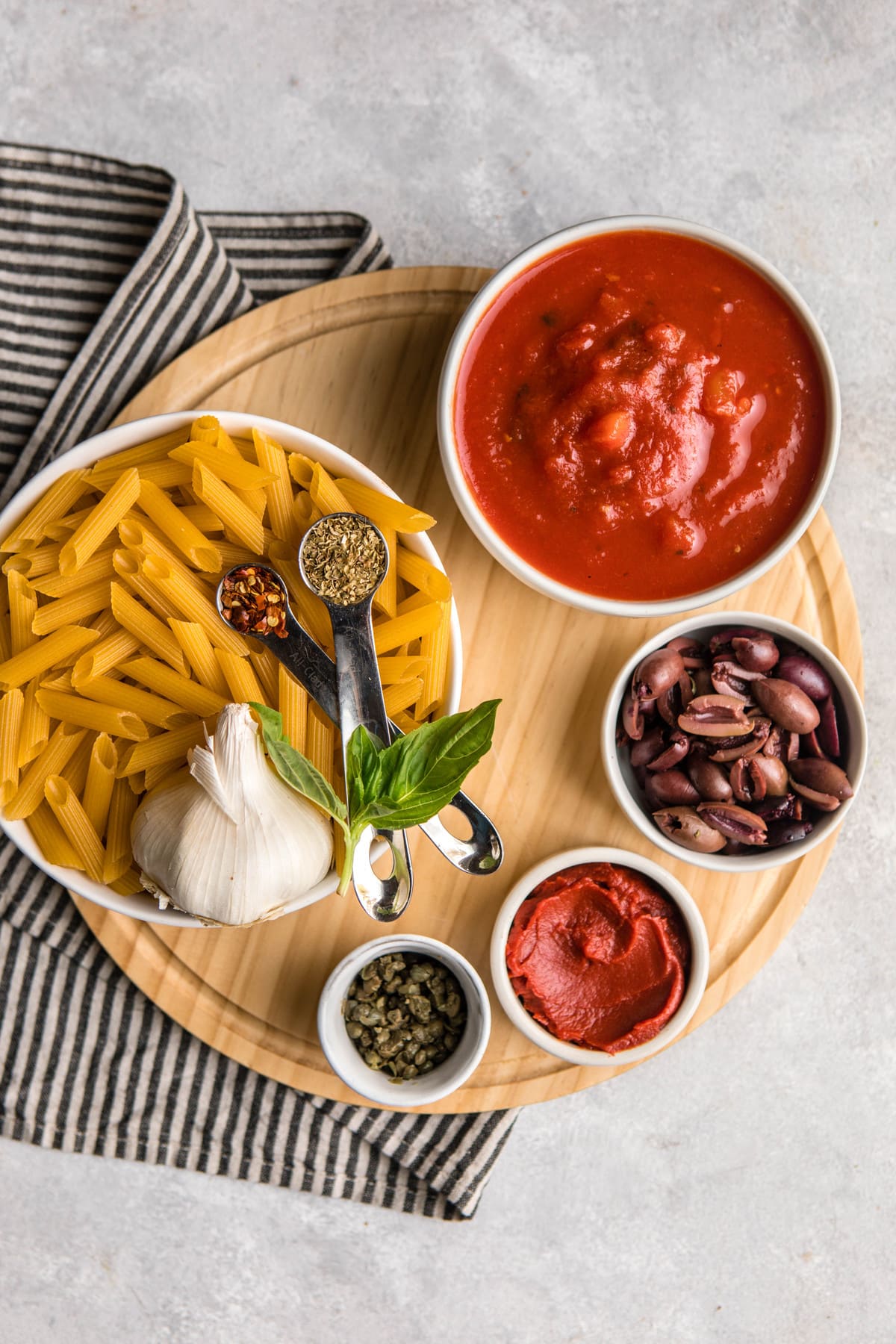 ingredients for penne puttanesca in small white cups on round wood cutting board
