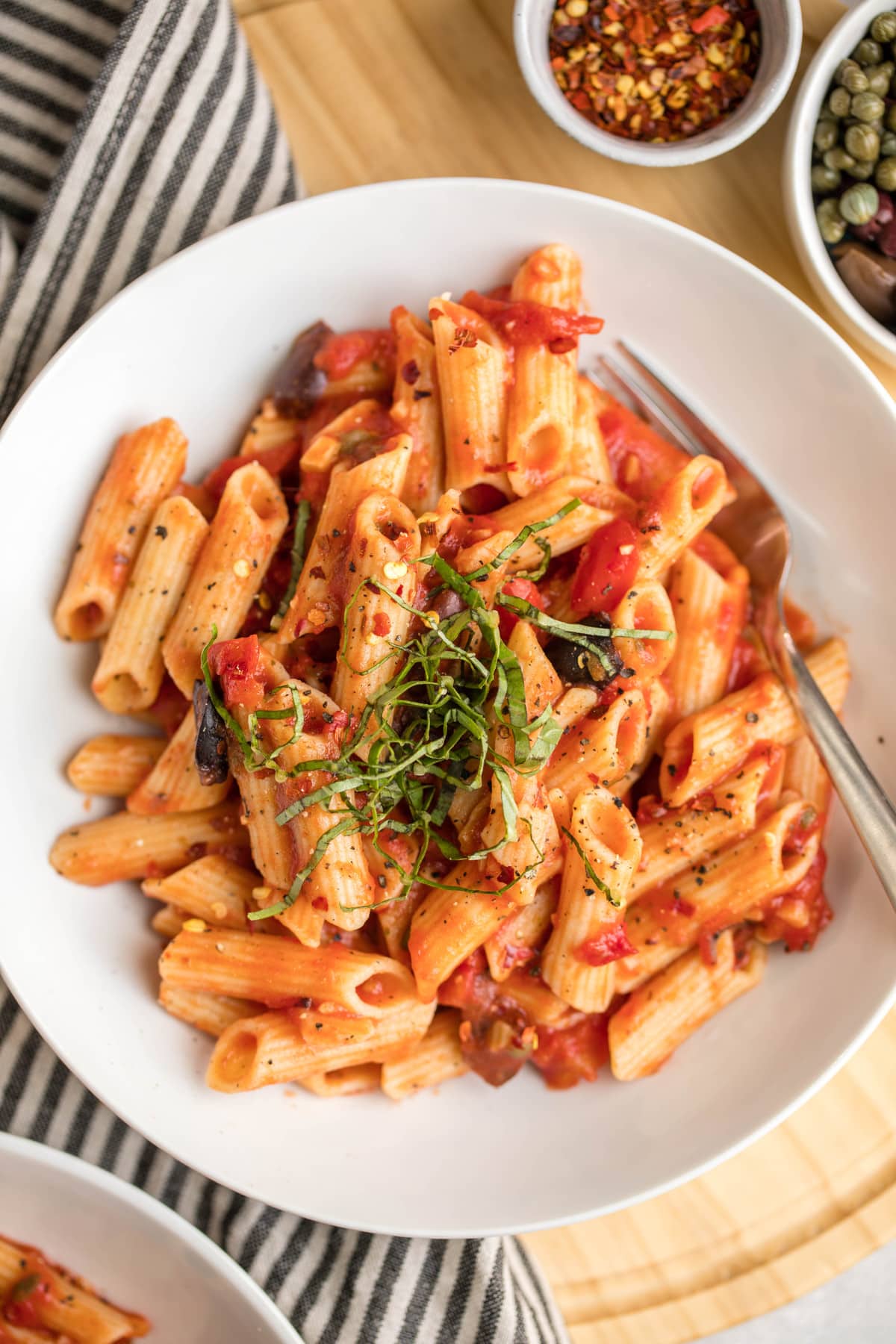 close up photo of penne puttanesca in white serving bowl topped with fresh basil