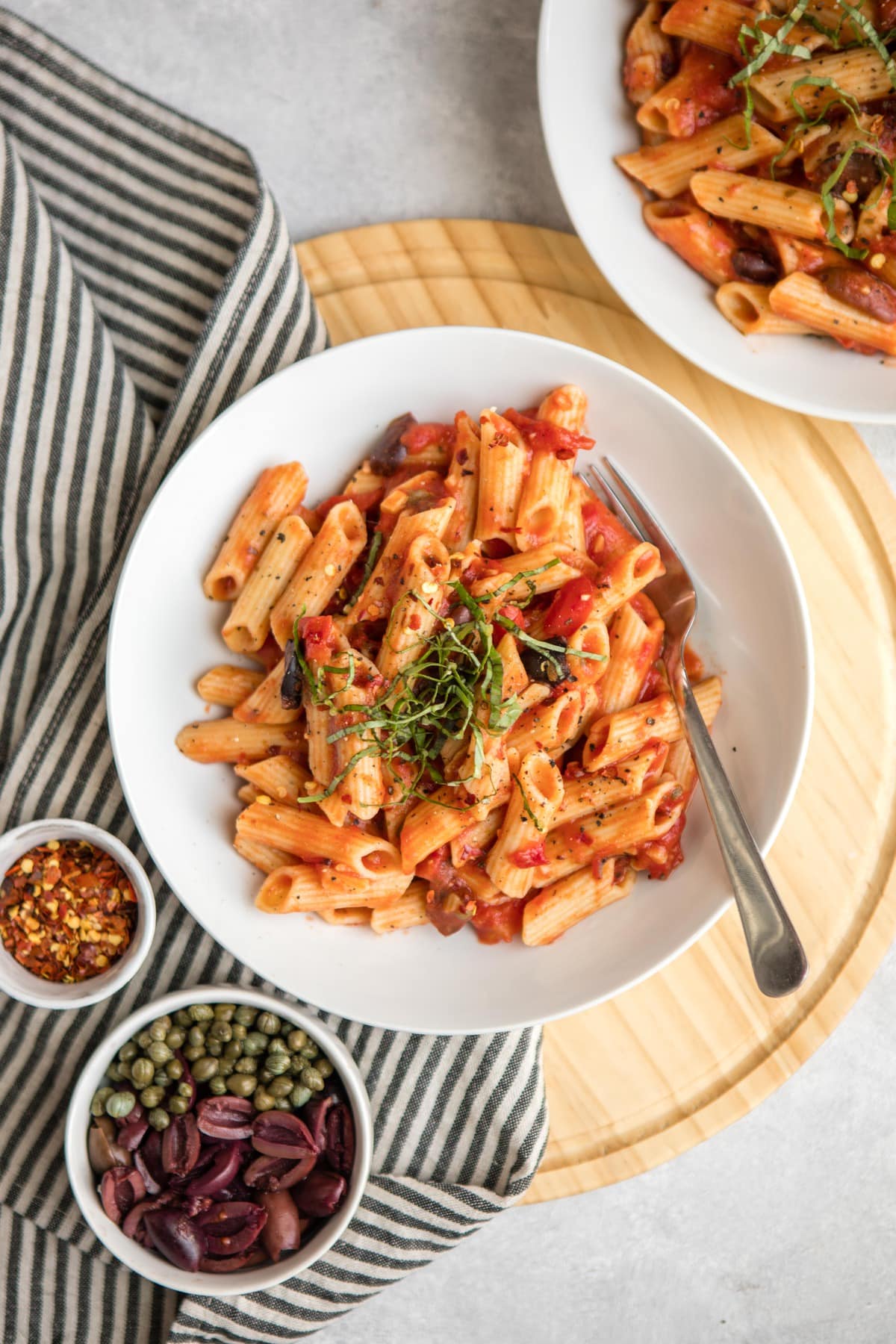 penne puttanesca topped with fresh basil and pepper in white pasta bowl on round wood cutting board