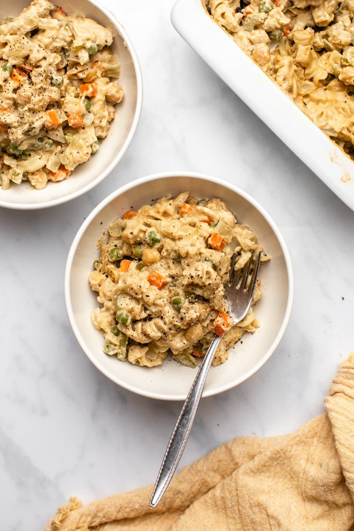 two white bowls of chickpea noodle casserole next to white casserole dish with yellow linen 