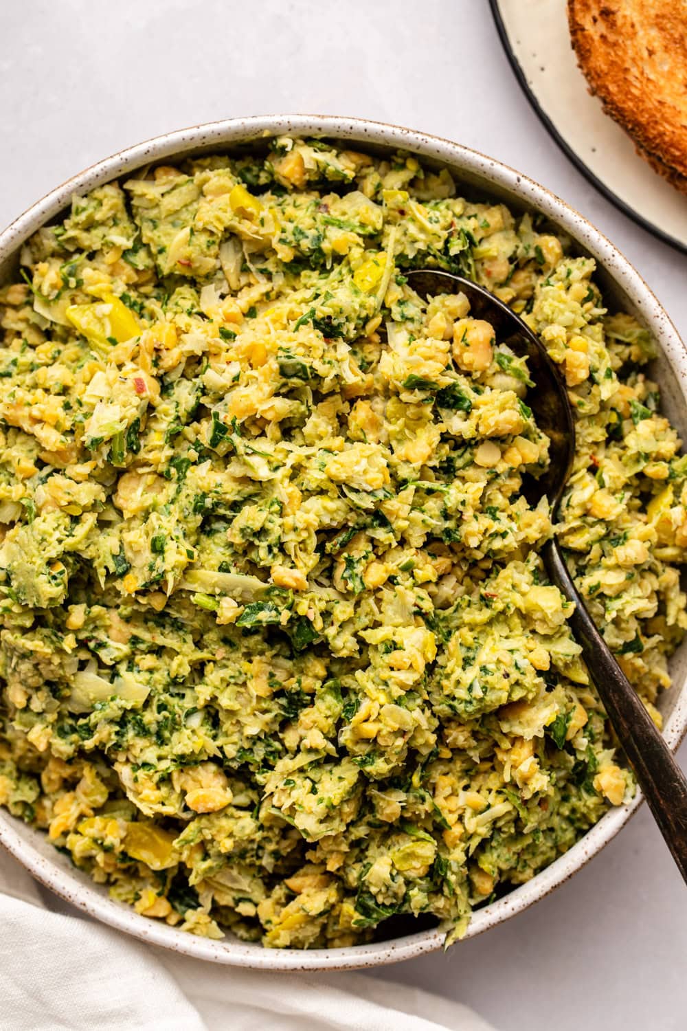 Spinach artichoke chickpea salad in white bowl with metal serving spoon