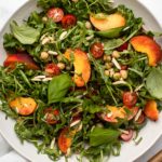 Peach, Arugula, & Chickpea Salad topped with grape tomatoes, basil, and almonds in a large white bowl with salad serving spoons off to the side