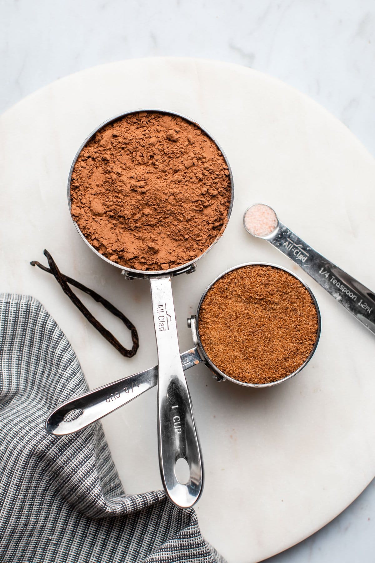 ingredients for hot chocolate mix in silver measuring cups on white marble serving board