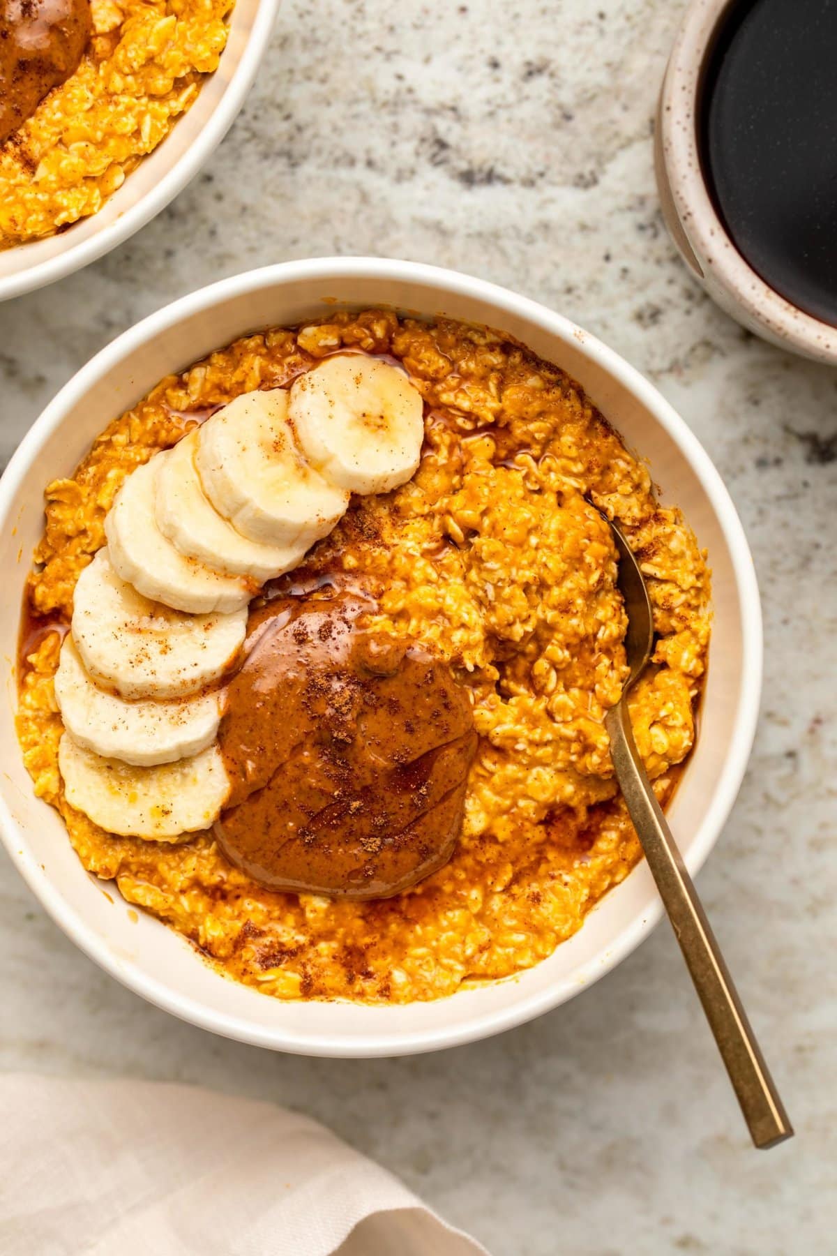Overhead photo of pumpkin spice latte overnight oats topped with banana, almond butter, and maple syrup with cup of coffee off to the side